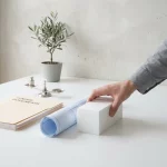 Architect legt rol blauwdruk en massamodel op witte desk naast aanbestedingsdossier en weegschaal, minimalistisch kantoor