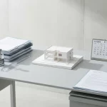 Wit architectuurmodel op minimalistisch bureau met documenten, datumstempel en kalender, in zacht ochtendlicht en strakke schaduwen