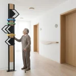 Richtingaanwijzer met kleurbanden in zorginstelling; oudere met wandelstok raakt bord aan in minimalistische gang