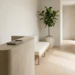 Minimalistische entree met receptiebalie, bank en sculpturale plant, lichte muren en houten vloer in rustig daglicht.