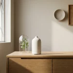 Minimalistische geur-diffuser van mat wit keramiek op licht eiken dressoir, met glazen stolp en plantje in ochtendlicht