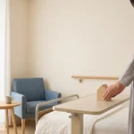 Verzorger houdt houten huisje op klinisch dienblad naast bed in warme zorgkamer met bedrail, handrail en daglicht
