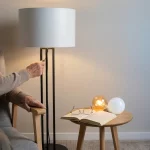Handen van oudere bij minimalistische vloerlamp, open boek en leesbril; warme en koele lichtbulb op bijzettafel.