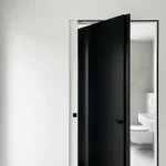 Matzwart minimalistisch interieurdeur in witte muur, op een kier naar moderne badkamer met witte wastafel en toilet