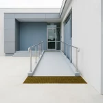 Rolstoeltoegankelijke entreehelling met handrail en tactiele strip naar moderne zorggebouwdeur in zacht daglicht