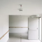 Sprinklerkop en rookmelder in wit plafond boven rustige gang van dementievriendelijk verpleeghuis met vluchtdeur en paniekbalk