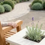 Oude hand op houten tuinbank naast verhoogde plantenbak met lavendel, minimalistische therapeutische tuin in ochtendlicht
