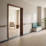 Rustige gang in verpleeghuis met brede deuropening naar lounge, handrail, contrastrand, zitnis en plant in daglicht.