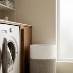 Open frontlader-wasmachine op hoogte in minimalistische wasruimte met wasmand, opgerolde handdoeken en zeepdispenser in zacht daglicht