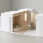 Minimalistisch architectuurmodel van zorggebouw met open deur, rustige gangzones, zitnis en tactiele wayfinding op grijze tafel