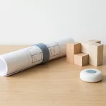 Opgerolde zorgkamer-plattegrond op houten bureau met modulaire houten blokken en verpleegoproepknop, minimalistisch licht