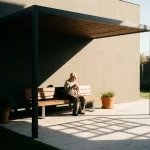 Oudere persoon met wandelstok op houten bank in zorgvriendelijke tuin, pergola schaduw en zonlicht op stenen terras