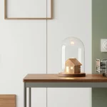 Houten miniatuurhuis onder glazen stolp op tafel in minimalistische zorgwoonkamer, naast gestapelde bedframes en bel