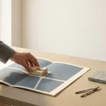 Architectenhand plaatst minimalistisch massingmodel op gevouwen sitetemap met stratenraster; kompas en materiaalstaal ernaast