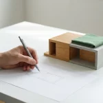 Architect tekent met potlood bouwschets naast houten en betonnen schaalmodel modern huis met groene materiaalstaal op dak