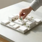 Hand plaatst laatste stuk in witte architectuurmaquette van nieuwe woonwijk met park en bomen op betonnen tafel