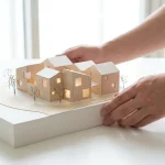 Architectonische maquette van woonblok met warm verlichte ramen, in handen op witte tafel, met bomen en wandelpad