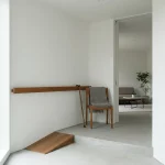 Handrail langs witte muur bij lage drempel met ramp, stoel met deken en wandelstok in minimalistische hal met open deur