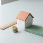 Wit modelhuis met opgetilde dakpan, euro munten en houten liniaal, groene isolatiepaneel op grijze achtergrond