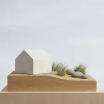 Minimalistisch modern wit huismodel op duinlandschap met gras en stenen, strak silhouet tegen bleke lucht in daglicht
