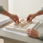 Schaalmodel van nieuwe woonwijk op tafel, handen plaatsen houten huisjes en bomen, blauwdruk en liniaal ernaast