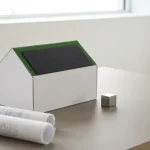 Wit modern huisarchitectuurmodel met zonnepanelen, blauwdrukken en metalen kubus voor bouwvoorschriften op neutrale tafel